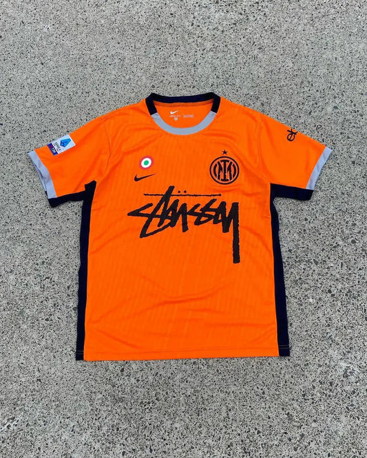 Orange Nike Inter Milan football jersey featuring black Stüssy logo and grey trim on concrete.